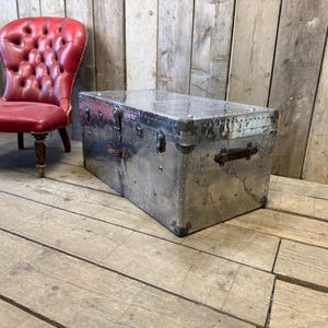 May include: A rectangular, silver metal trunk with riveted construction and leather handles. The trunk has a reflective surface and is secured with metal clasps. A red leather chair is visible in the background.