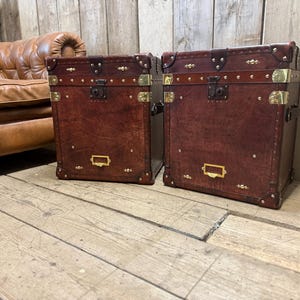 Puede incluir: Dos baúles de almacenamiento a juego, de cuero marrón oscuro con herrajes de latón. Los baúles tienen forma rectangular con esquinas redondeadas, detalles de latón y una pequeña placa rectangular de latón. Aproximadamente 60 cm de altura.