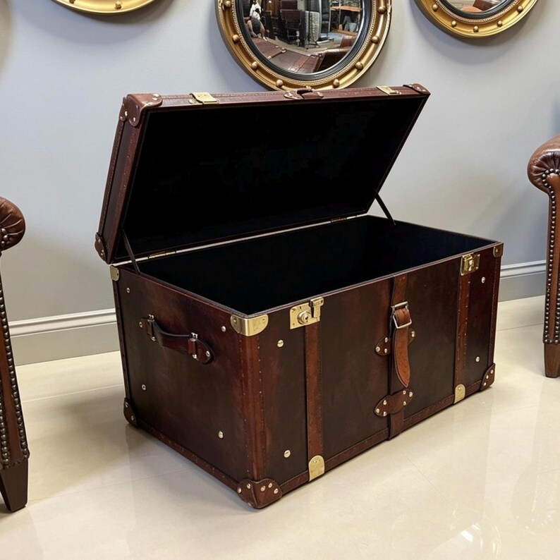 Handmade dark brown leather steamer trunk with brass corner plates, stud trim and leather carry straps, photographed between two leather armchairs