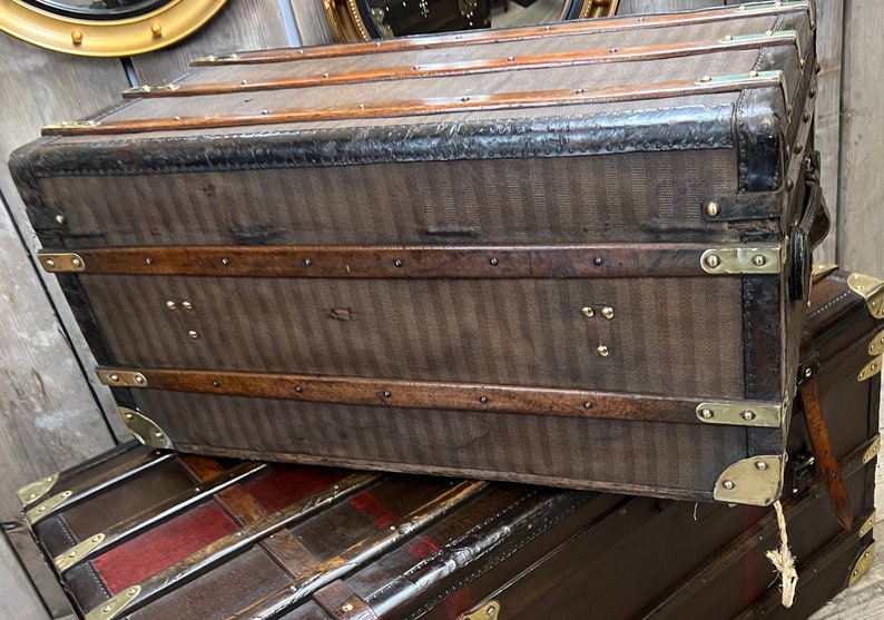 Puede incluir: Ba&uacute;l marr&oacute;n antiguo con patr&oacute;n de rayas verticales, ribetes de cuero y herrajes de lat&oacute;n. El ba&uacute;l es rectangular con una parte superior curva y est&aacute; apilado sobre otro ba&uacute;l. Art&iacute;culos de almacenamiento de viaje vintage.