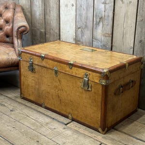 May include: A large, rectangular wooden trunk with a light brown finish. The trunk features brass hardware, including corner protectors, latches, and a small rectangular plate on the top. Leather handles are on the sides. The trunk is a vintage style.