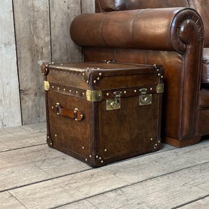 May include: A square brown leather trunk featuring brass corner reinforcements, brass studs along the edges, and two brass locking latches on the front. The trunk includes a brown leather side handle and is positioned next to a brown leather sofa.