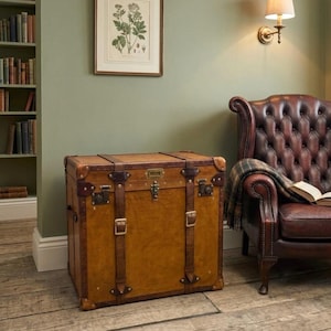 Puede incluir: Un baúl rectangular de estilo vintage, de color marrón claro, con correas y detalles de cuero marrón oscuro. El baúl tiene herrajes metálicos y una cerradura. La escena incluye un sillón de cuero y una estantería.