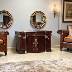 Handmade dark brown leather steamer trunk with brass corner plates, stud trim and leather carry straps, photographed between two leather armchairs