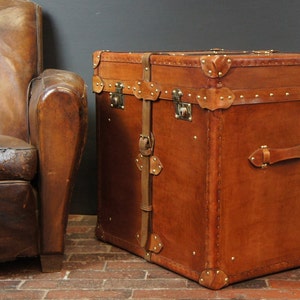 May include: A brown leather trunk with brass hardware and straps. The trunk is closed and has a handle on the front.