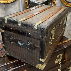 Puede incluir: Un ba&uacute;l marr&oacute;n vintage con herrajes de lat&oacute;n y detalles de cuero. El ba&uacute;l presenta un patr&oacute;n de rayas en el exterior y listones de madera en la parte superior. El ba&uacute;l rectangular tiene un asa en el lateral.