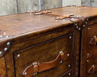 Puede incluir: Primer plano de dos baúles de madera marrón con asas y detalles de cuero. Los baúles tienen tachuelas metálicas a lo largo de los bordes y correas de cuero en la parte superior. La madera tiene un tono rico y cálido, y el diseño evoca una estética vintage.