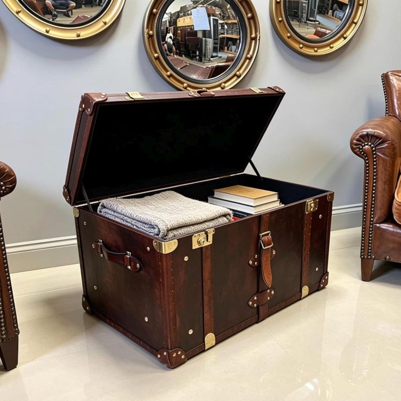 Handmade dark brown leather steamer trunk with brass corner plates, stud trim and leather carry straps, photographed between two leather armchairs