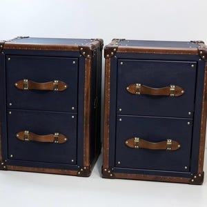 May include: Two matching navy blue two-drawer storage cabinets featuring brown leather-like trim, brass-colored metal corner reinforcements, and brown leather-like drawer pulls secured with brass-colored rivets on each drawer front.