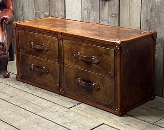Puede incluir: Un aparador de cuero marrón con cuatro cajones y herrajes de latón. El aparador es rectangular y tiene un estilo vintage, tipo maleta de viaje. Los cajones tienen tiradores de cuero y están enmarcados con protectores de esquina de latón.