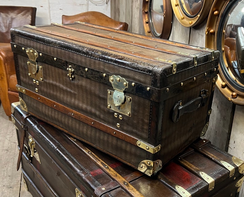 Puede incluir: Un ba&uacute;l marr&oacute;n vintage con herrajes de lat&oacute;n, que incluyen protectores de esquinas, pestillos y una cerradura central. El ba&uacute;l presenta un exterior estampado y detalles de madera. Est&aacute; apilado encima de otro ba&uacute;l similar.