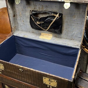 Puede incluir: Un ba&uacute;l antiguo marr&oacute;n abierto, con herrajes de lat&oacute;n, que revela un interior azul y un forro a rayas. El ba&uacute;l tiene forma rectangular con protectores de esquina met&aacute;licos y un peque&ntilde;o bolsillo de almacenamiento.