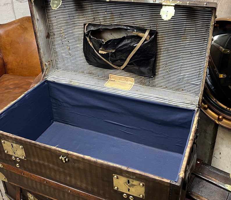 Puede incluir: Un ba&uacute;l antiguo marr&oacute;n abierto, con herrajes de lat&oacute;n, que revela un interior azul y un forro a rayas. El ba&uacute;l tiene forma rectangular con protectores de esquina met&aacute;licos y un peque&ntilde;o bolsillo de almacenamiento.