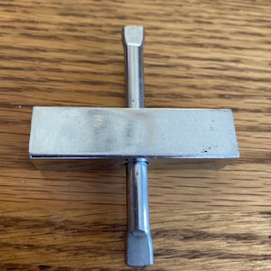 May include: A silver metal tool with a rectangular base and a long, thin, pointed shaft extending from the center of the base.