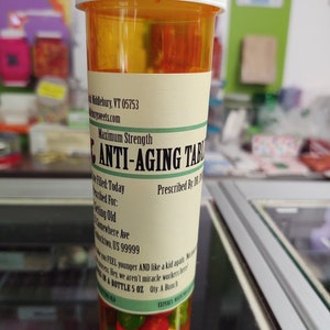 May include: A clear plastic bottle with a white lid and a yellow label that reads "Maximum Strength ANTI-AGING TABLETS Prescribed By: Dr. Time". The label also includes the text "Filet Today", "United For: Getting Old", "Somewhere Ave", "Blacktwo, US 99999", "Make you FEEL younger AND like a kid again. Not sure about that. Hey, we aren't miracle workers here!" and "IN A BOTTLE 5 OZ Qty: A Bunch". The bottle is filled with colorful candy.