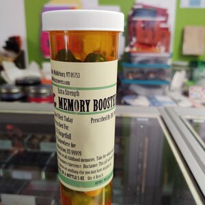 May include: A clear plastic bottle with a white lid filled with green pills. The label reads "Extra Strength Memory Booster" and includes instructions for use. The bottle is labelled "Prescribed By: Dr. S. Forgetfull" and "City: A Bunch".
