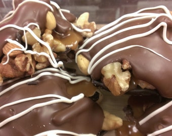 May include: Close-up of chocolate-covered caramel clusters with nuts and white chocolate drizzle. The clusters are arranged on a surface.