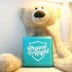 May include: A teal blue wooden sign with a white shield shape and the text "Choose the Right" in a white vintage font. The sign is sitting in front of a beige teddy bear.