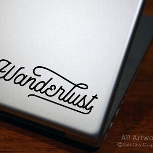 May include: A silver laptop with a black vinyl sticker that says "Wanderlust" in a cursive font.