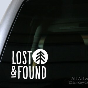 May include: White vinyl decal with the text "Lost & Found" and a stylized tree graphic. The decal is applied to the rear window of a car.