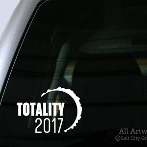 May include: A white vinyl sticker with a crescent moon shape and the text "Totality 2017". The sticker is on the back window of a car.
