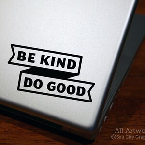 Photo of Salt City Graphics Be Kind Do Good vinyl decal in Black, in a block typeface with the phrase inside a banner; decal is shown on the bottom right corner of a silver laptop lid atop a dark wood surface. Photo and artwork copyright Salt City Graphics®.