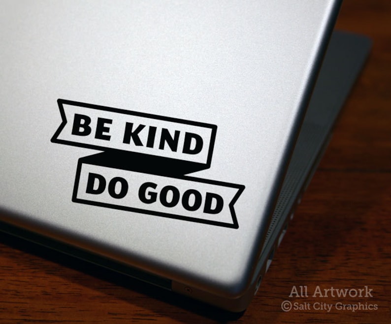 Photo of Salt City Graphics Be Kind Do Good vinyl decal in Black, in a block typeface with the phrase inside a banner; decal is shown on the bottom right corner of a silver laptop lid atop a dark wood surface. Photo and artwork copyright Salt City Graphics®.