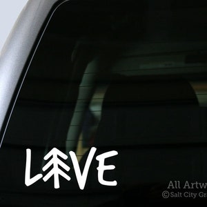 May include: White vinyl decal with the word "LOVE" with a stylized pine tree replacing the "O". The decal is on the back window of a car.