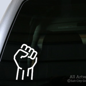 May include: White vinyl sticker of a raised fist with the fingers closed. The sticker is on the back window of a car.