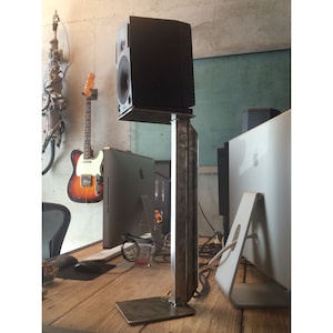 Puede incluir: Un altavoz negro sobre un soporte de metal. El soporte es de metal y tiene una base cuadrada. El altavoz está sobre una mesa de madera.