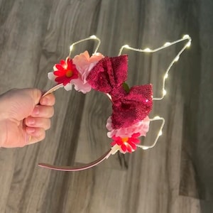 以下が含まれることがあります： ピンク色のヘアバンド。スパンコール付きのバーガンディ色のリボン、ピンクと赤の花、小さな白いライトで飾られています。ヘアバンドは木製の背景に対して保持されています。