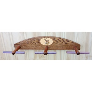 May include: A wooden wall-mounted gun rack with three hooks and a deer silhouette design. The rack is made of dark wood with a light wood inlay. The hooks are made of dark wood with purple rubber grips.