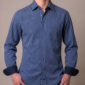 May include: A long-sleeve button-down shirt featuring vertical stripes in alternating shades of navy blue and light blue. The shirt includes a standard collar, a single chest pocket, and dark cuffs with a subtle, small-scale geometric pattern.