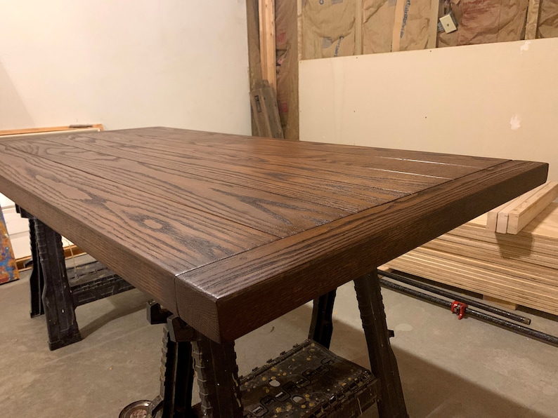 Puede incluir: Una tapa de mesa de madera marr&oacute;n oscuro con un borde grueso biselado. La tapa de la mesa est&aacute; hecha de varias tablas de madera que se han unido. La veta de la madera es visible y la tapa de la mesa tiene un acabado liso y pulido.