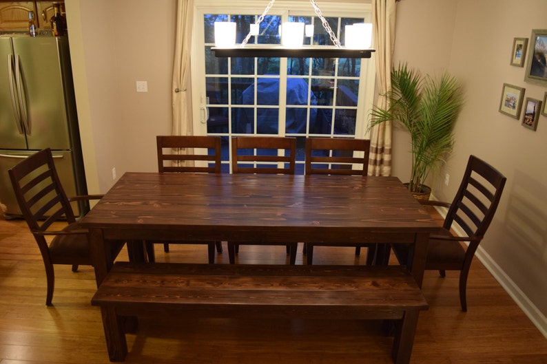 Puede incluir: Una mesa de comedor de madera marr&oacute;n oscuro con seis sillas y un banco. La mesa tiene un acabado r&uacute;stico y est&aacute; hecha de madera maciza. Las sillas tambi&eacute;n son de madera y tienen un dise&ntilde;o sencillo. El banco tambi&eacute;n es de madera y tiene un acabado similar al de la mesa.