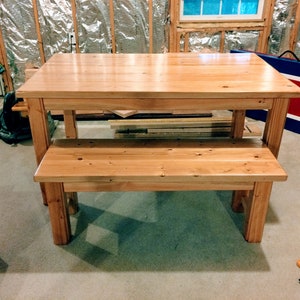 Puede incluir: Un conjunto de mesa y banco de madera natural. La mesa tiene una tapa rectangular y cuatro patas. El banco tiene un asiento rectangular y cuatro patas. El conjunto est&aacute; en una habitaci&oacute;n con suelo de moqueta.