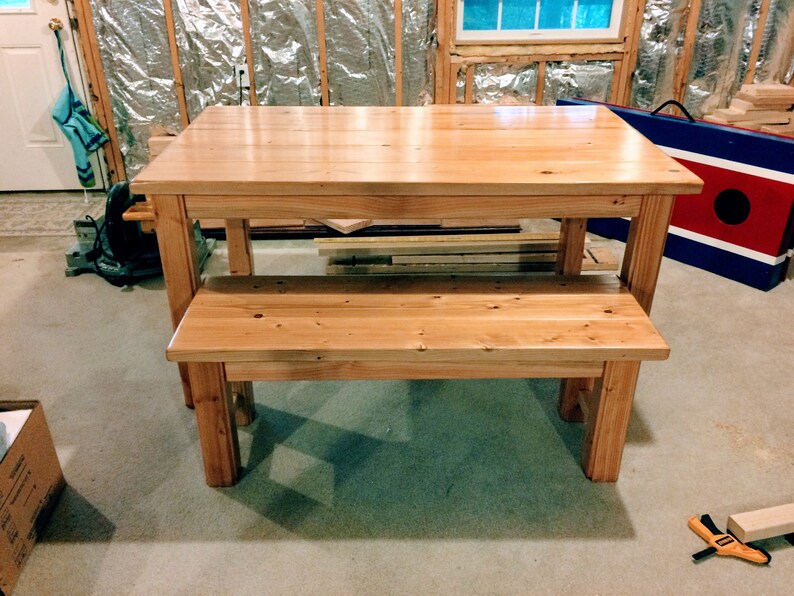 Puede incluir: Un conjunto de mesa y banco de madera natural. La mesa tiene una tapa rectangular y cuatro patas. El banco tiene un asiento rectangular y cuatro patas. El conjunto est&aacute; en una habitaci&oacute;n con suelo de moqueta.