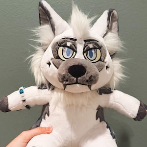 May include: A plush wolf toy featuring white and gray fur, large expressive blue eyes, pointed ears, and a small beaded bracelet on one arm. The creature has dark gray paw pads with light-colored claws and a tuft of white fur on its head.