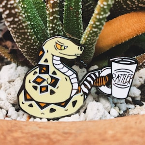 May include: A yellow and black enamel pin of a rattlesnake coiled up with a white coffee cup that says "RATTLESNAKE" in black lettering.
