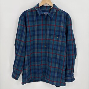 Puede incluir: Una camisa de cuadros de manga larga en tonos azules, verde azulado y rojo. La camisa tiene un cuello con cremallera, un bolsillo en el pecho y puños abotonados. La camisa está colgada en una percha de madera.