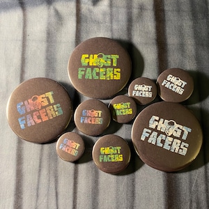 Könnte beinhalten: Eine Sammlung von acht runden schwarzen Knöpfen mit dem Text "Ghost Facers" in einem Regenbogen-Farbverlauf. Die Knöpfe sind in verschiedenen Größen.