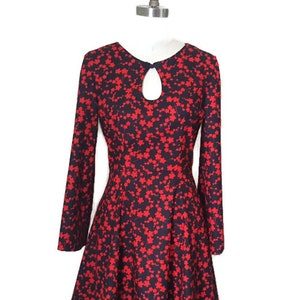 K&ouml;nnte beinhalten: Ein schwarzes Kleid mit rotem Blumendruck, langen &Auml;rmeln und einem Schl&uuml;ssellochausschnitt. Das Kleid hat einen taillierten Oberteil und einen ausgestellten Rock.