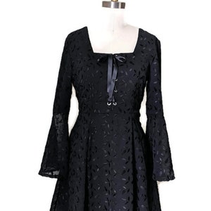 Pode incluir: Um vestido preto com decote quadrado e mangas compridas. O vestido tem um detalhe de renda na frente e um la&ccedil;o de fita no pesco&ccedil;o. O vestido &eacute; feito de um tecido leve com um padr&atilde;o de folhas.