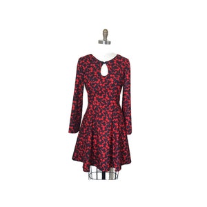 K&ouml;nnte beinhalten: Ein rotes und schwarzes Blumenkleid mit langen &Auml;rmeln und einem Schl&uuml;ssellochausschnitt. Das Kleid hat ein tailliertes Oberteil und einen ausgestellten Rock.