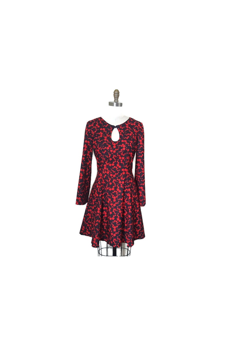 K&ouml;nnte beinhalten: Ein rotes und schwarzes Blumenkleid mit langen &Auml;rmeln und einem Schl&uuml;ssellochausschnitt. Das Kleid hat ein tailliertes Oberteil und einen ausgestellten Rock.