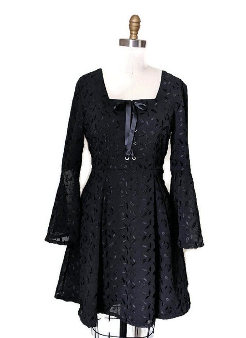 Pode incluir: Um vestido preto com decote quadrado e mangas compridas. O vestido tem um detalhe de renda na frente e um la&ccedil;o de fita no pesco&ccedil;o. O vestido &eacute; feito de um tecido leve com um padr&atilde;o de folhas.
