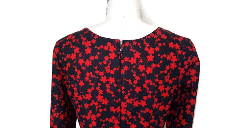 K&ouml;nnte beinhalten: Ein schwarzes Kleid mit rotem Blumendruck. Das Kleid hat einen runden Ausschnitt und lange &Auml;rmel. Der R&uuml;cken des Kleides hat einen Rei&szlig;verschluss.