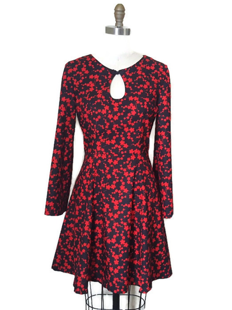 K&ouml;nnte beinhalten: Ein schwarzes Kleid mit rotem Blumendruck, langen &Auml;rmeln und einem Schl&uuml;ssellochausschnitt. Das Kleid hat einen taillierten Oberteil und einen ausgestellten Rock.