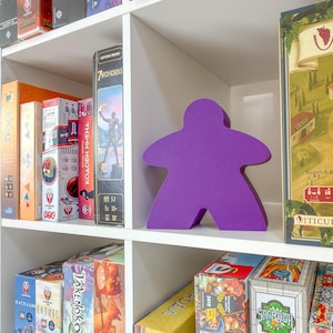 Op de afbeelding: Een witte boekenplank vol bordspellen. Een groot, paars spelstuk valt op. Speldozen met titels als "Dune", "Viticulture" en "The Castles of Burgundy". De plank is georganiseerd met diverse speldozen, met kleurrijke illustraties.