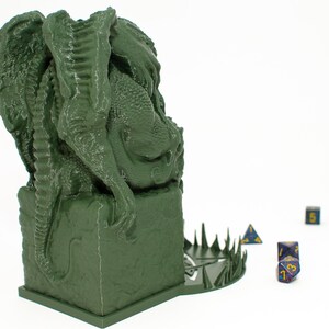 May include: Green 3D printed dragon statue with a base that has a circular design with spikes around the edge. The dragon is sitting on a square base. There are several blue dice on the table in front of the dragon.